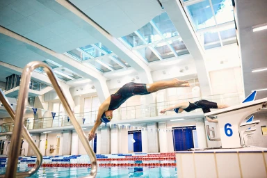 Swimmers in racing swimsuits diving into an indoor swimming pool from starting blocks under a glass skylight ceiling.