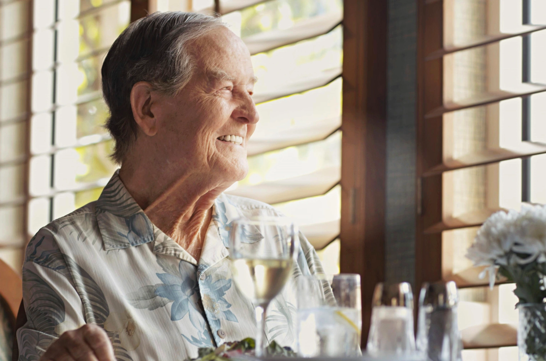 Elderly man wearing a floral patterned short-sleeve shirt smiling while seated near a window with wooden blinds.