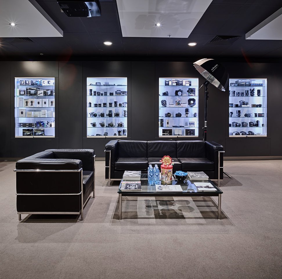 A showroom featuring camera equipment in glass cabinets, black leather seating, and a modern interior design with neutral carpet.