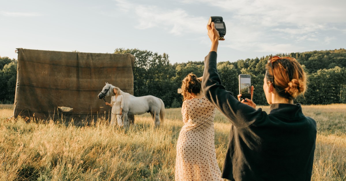 Outdoor photo shoot with a white horse and two adult women using professional camera equipment in a grassy field with trees in the background.