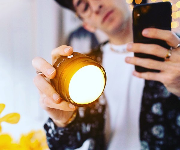 Person holds illuminated brown cylinder in hand, using a smartphone to capture a picture. Soft lighting and blurred background.