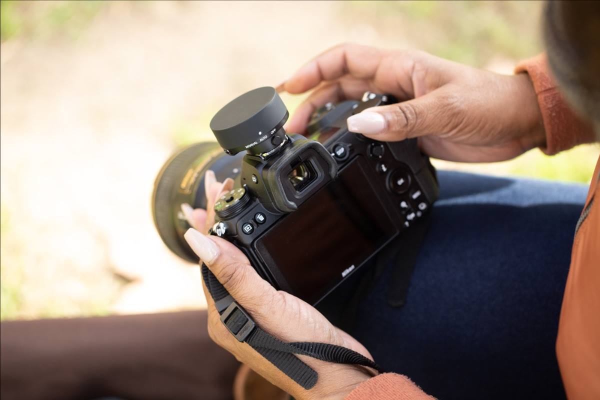 Close-up of a Black woman holding a Nikon mirrorless camera with a circular external viewfinder attachment in an outdoor setting.