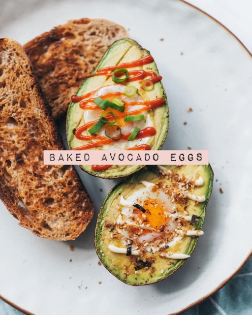 Overhead shot of a plate with two avocado halves filled with baked eggs, garnished with green onions and a drizzle of sauce, alongside toast.