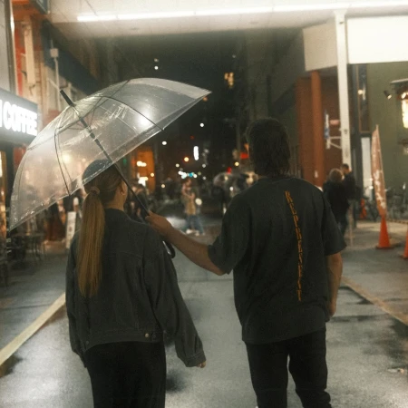 Couple walking in the city at night, sharing a clear umbrella under the street lights, with wet pavement and pedestrians around.