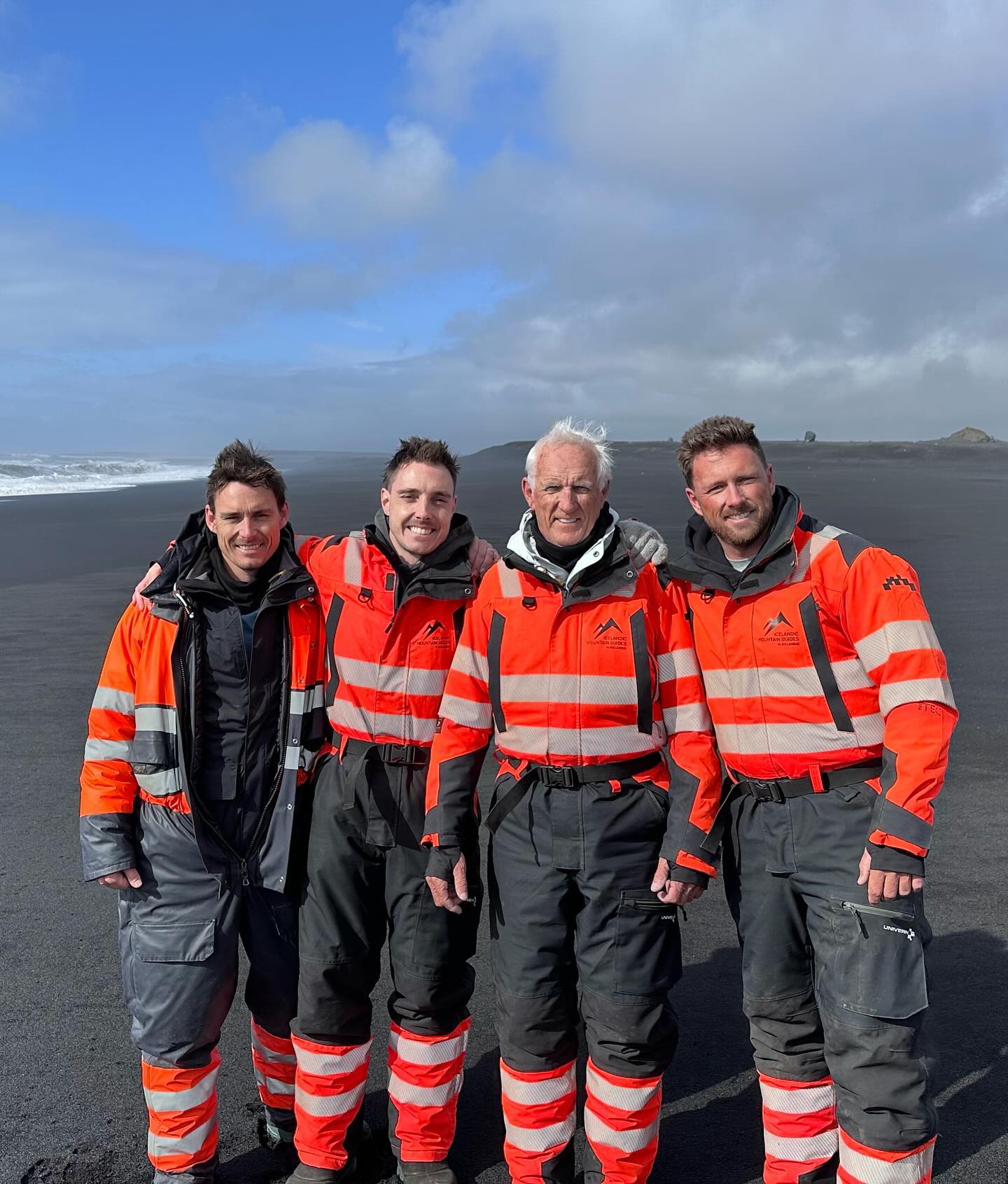 A group of four people in safety gear posing on a black sand beach under a partly cloudy sky, with ocean waves in the background.
