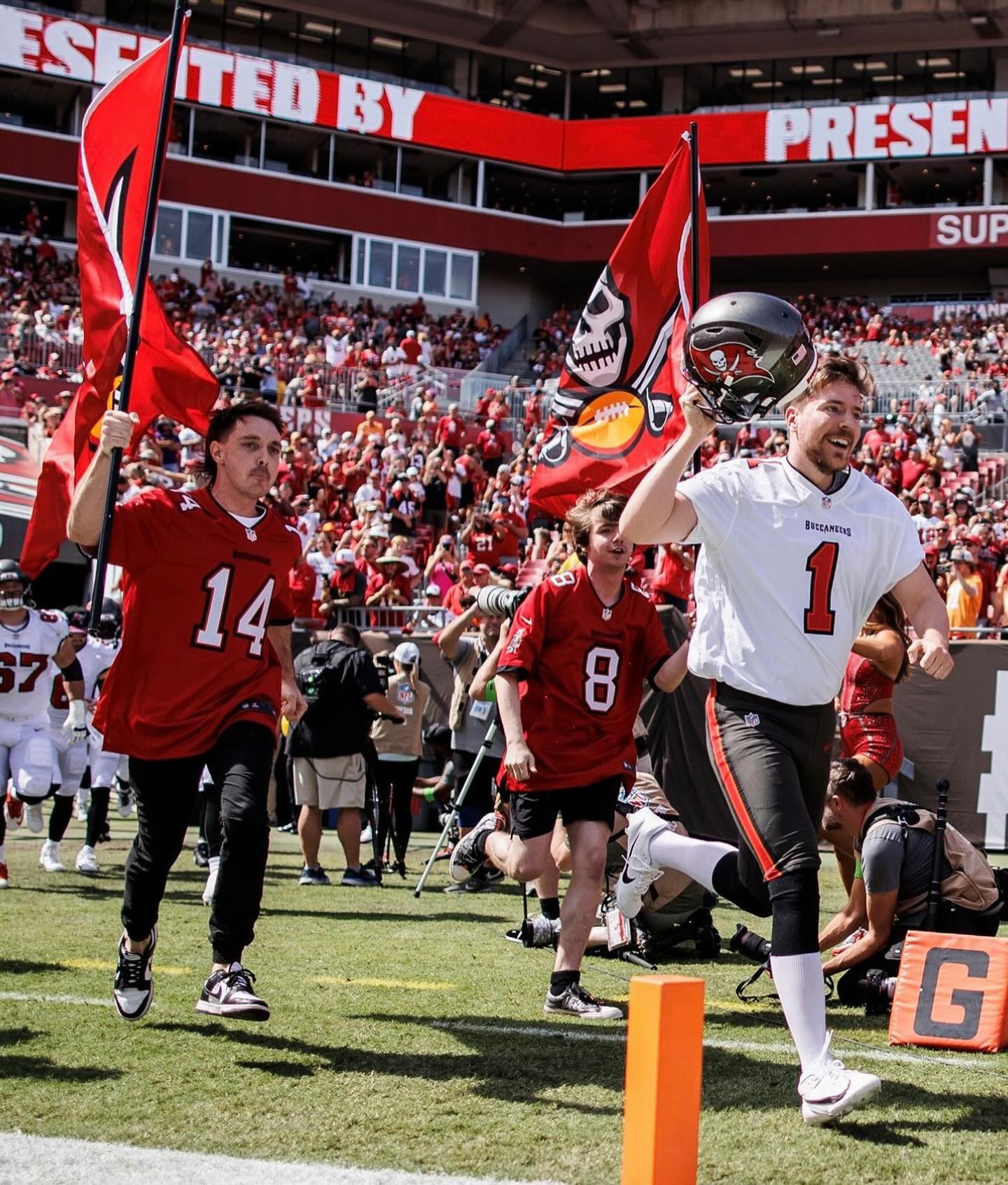 Three people in football jerseys run onto a stadium field carrying flags, with a crowd in the background on a sunny day.