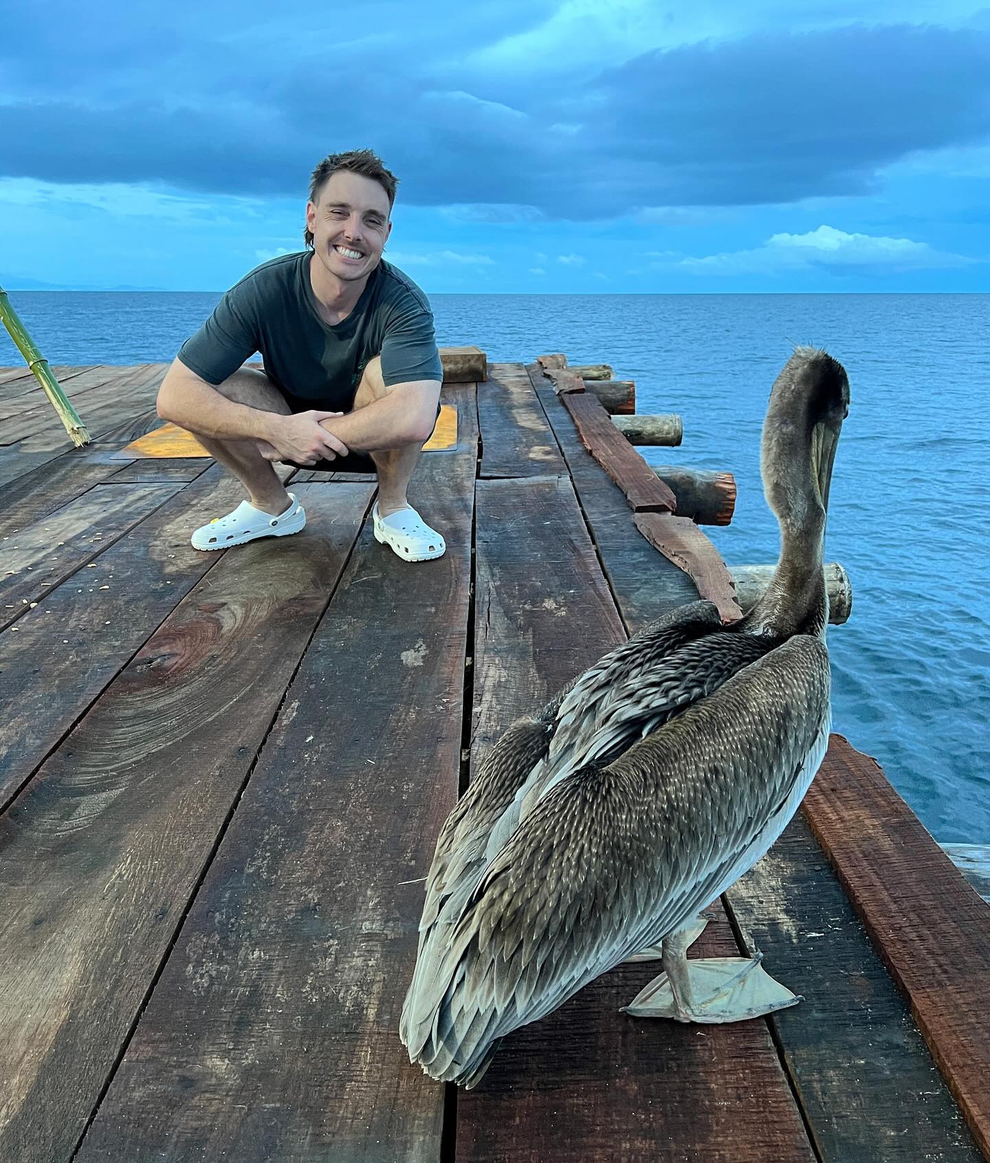 Adult man crouches on a dock near a large pelican bird overlooking the ocean on a cloudy day with grey-blue sky at daytime.