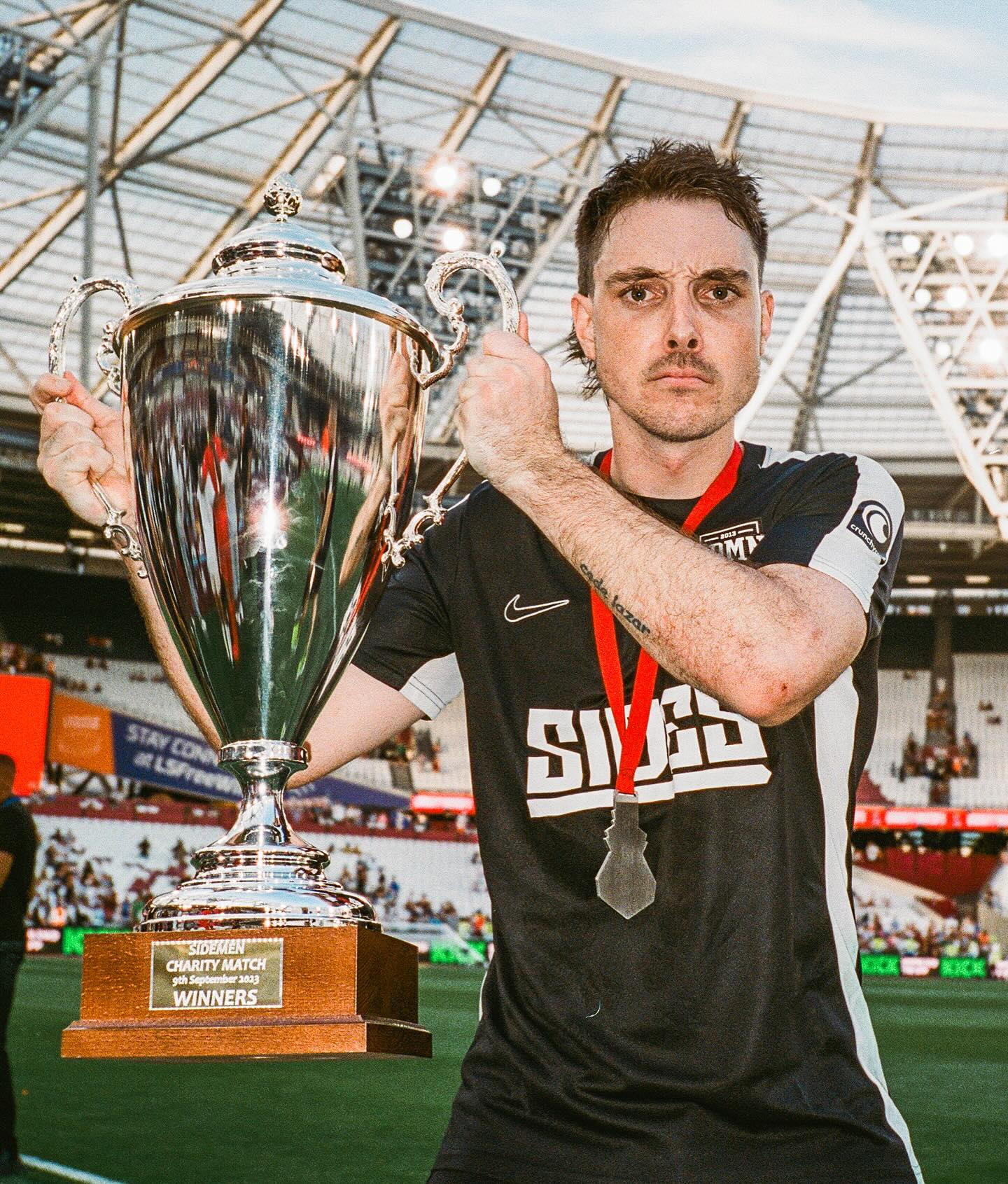 Man in sportswear holds a trophy with a red medal in an outdoor stadium, capturing a celebratory moment. 