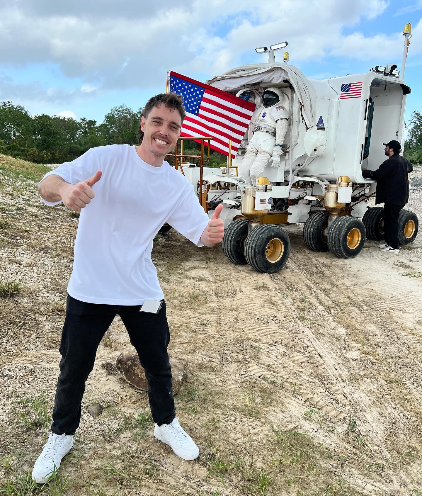 Smiling man poses in front of a lunar rover with an astronaut and an American flag in a dirt outdoor environment on a sunny day.