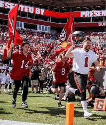 Group of men in football jerseys running onto a stadium field, holding team flags and a helmet under bright daylight.