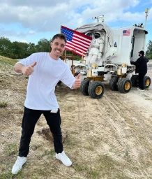 Content creator LazarBeam smiling and giving thumbs up in front of a white rover vehicle with an American flag and spacesuit.