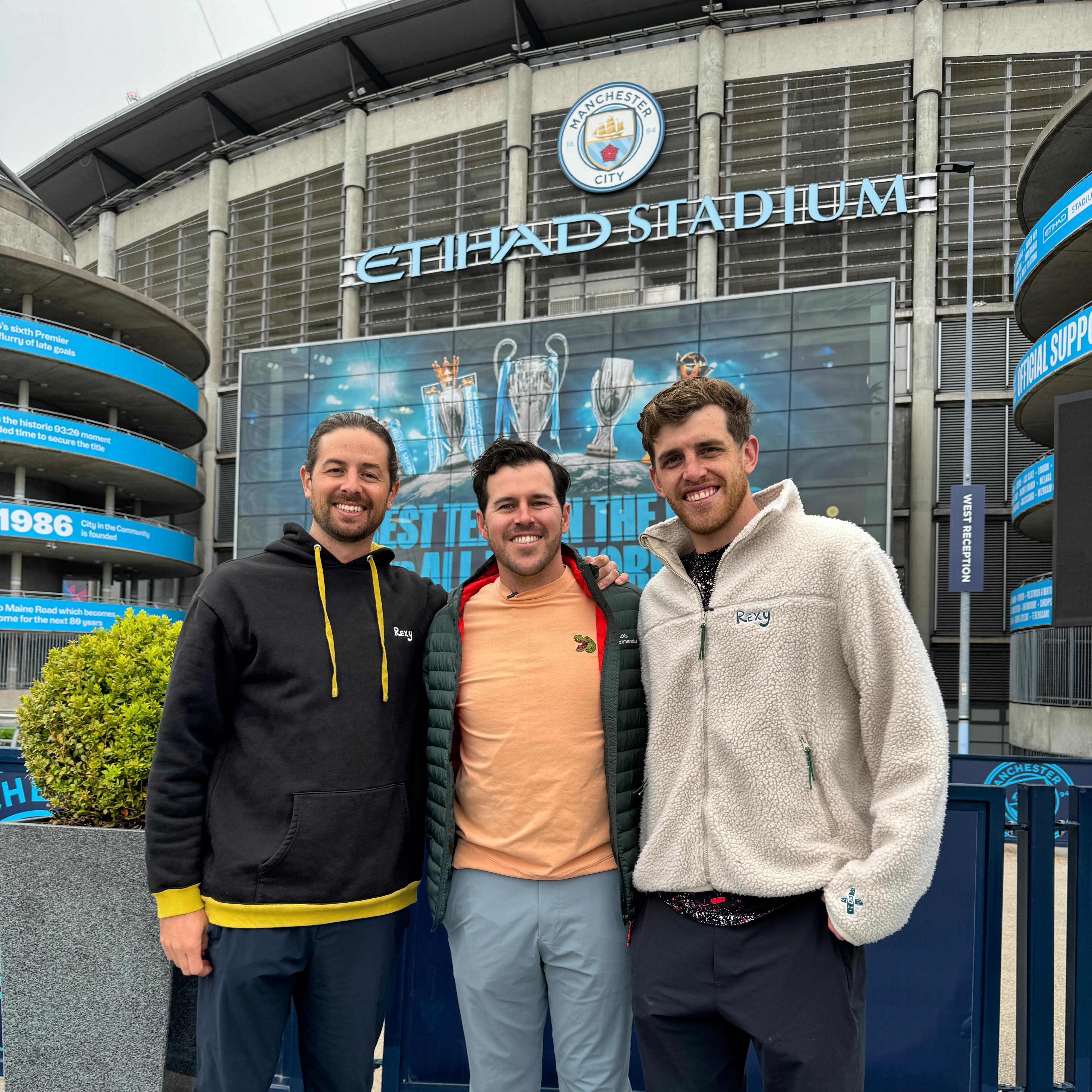 Three adult men pose in front of Etihad Stadium, home of Manchester City, wearing casual clothing. Stadium and cloudy sky visible.