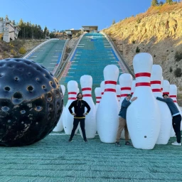 Giant inflatable bowling pins and a massive black bowling ball set up at the base of a large outdoor ski jump ramp.