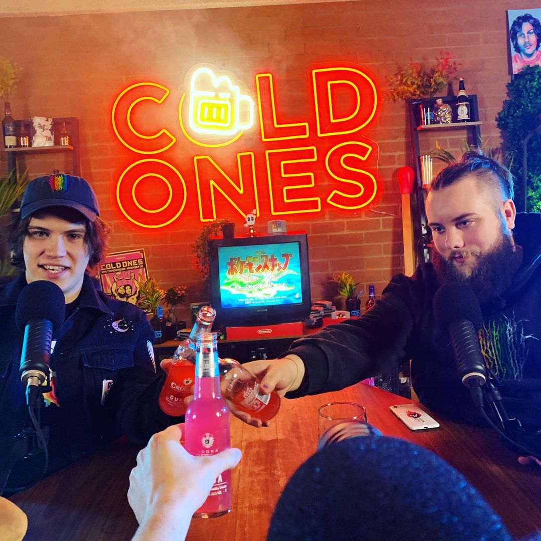 Two men sitting at a table with microphones, holding up pink and red bottles, with a neon sign in the background.