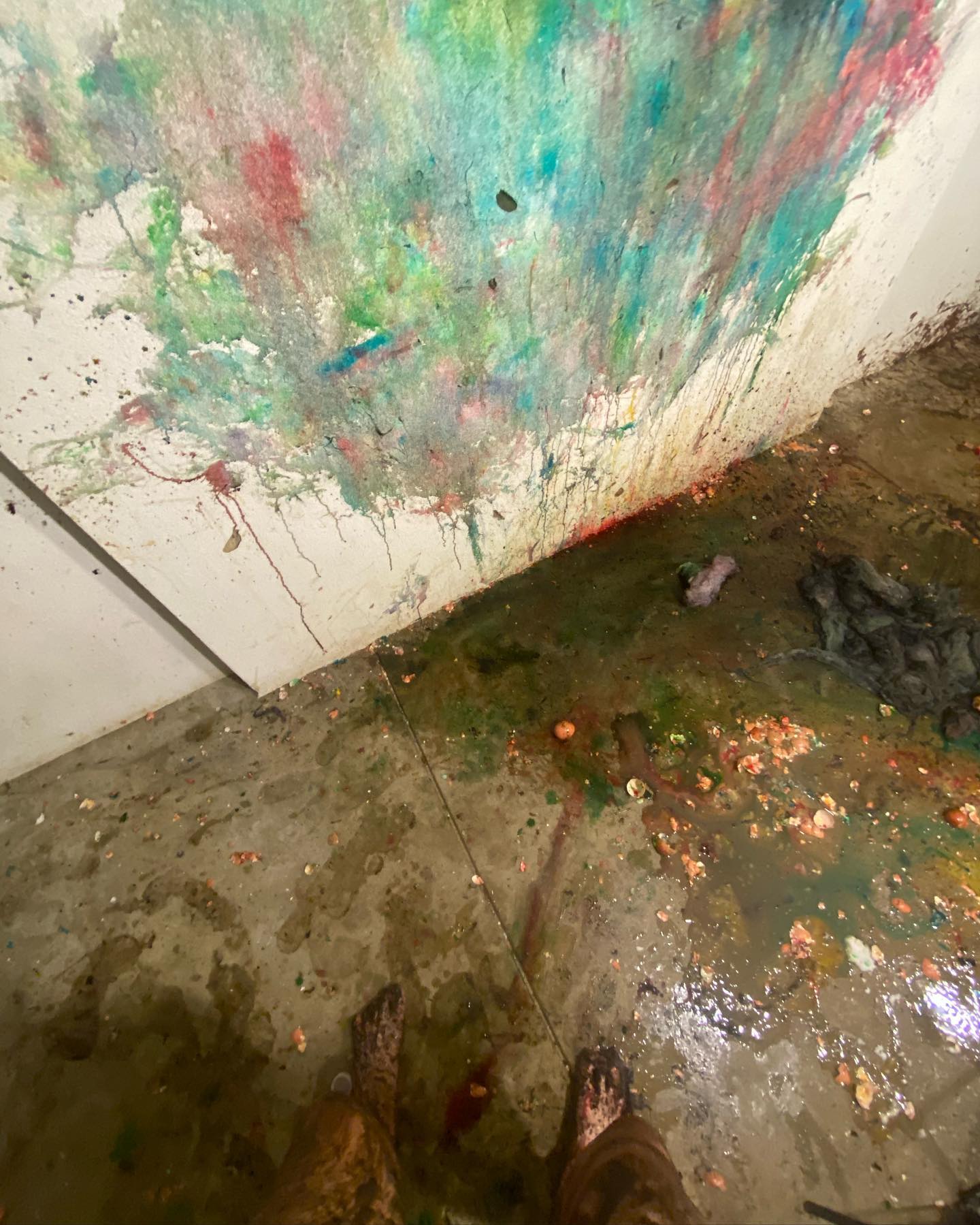 High-angle shot of a person's feet standing in a room with paint-splattered walls and floors, creating an abstract, colorful, messy scene.