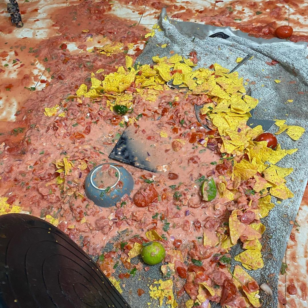 Overhead view of a messy floor with spilled salsa, tortilla chips, limes, tomatoes, and a broken bowl on a towel. 
