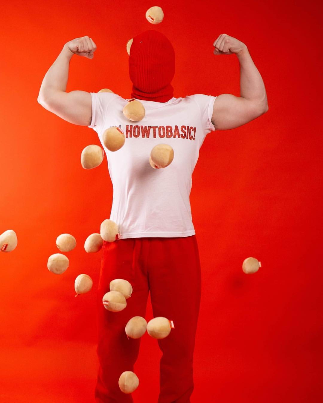 Muscular adult in a red balaclava and sweats posing with eggs against a red backdrop, showcasing strength and humor in studio lighting.