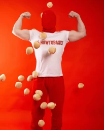 Person in red balaclava and t-shirt flexing muscles amidst falling potatoes against red background
