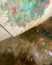 First-person overhead view of bare feet standing on a messy concrete floor covered in colorful splatters, food debris, and a painted wall.