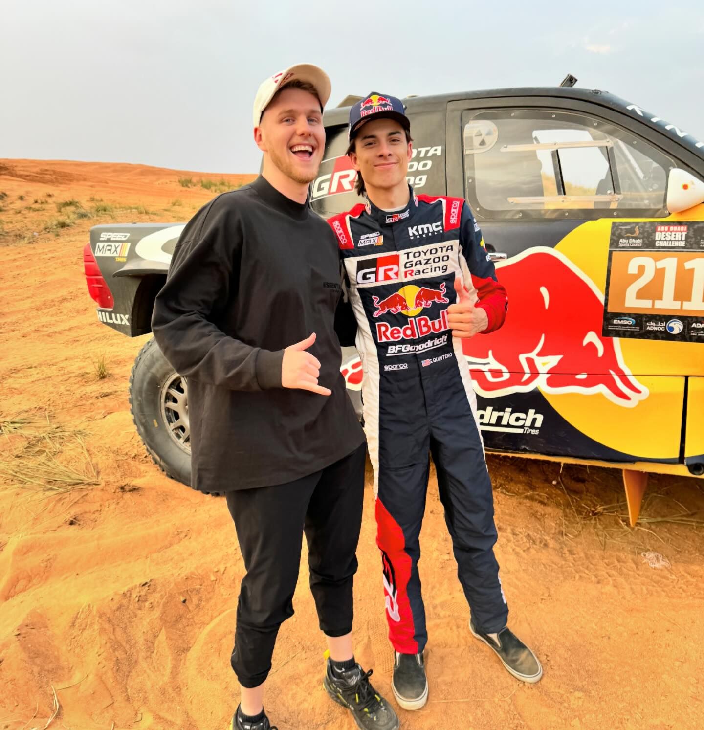 Two men pose in front of a Dakar Rally car in a desert landscape under a cloudy sky. One man wears racing attire.