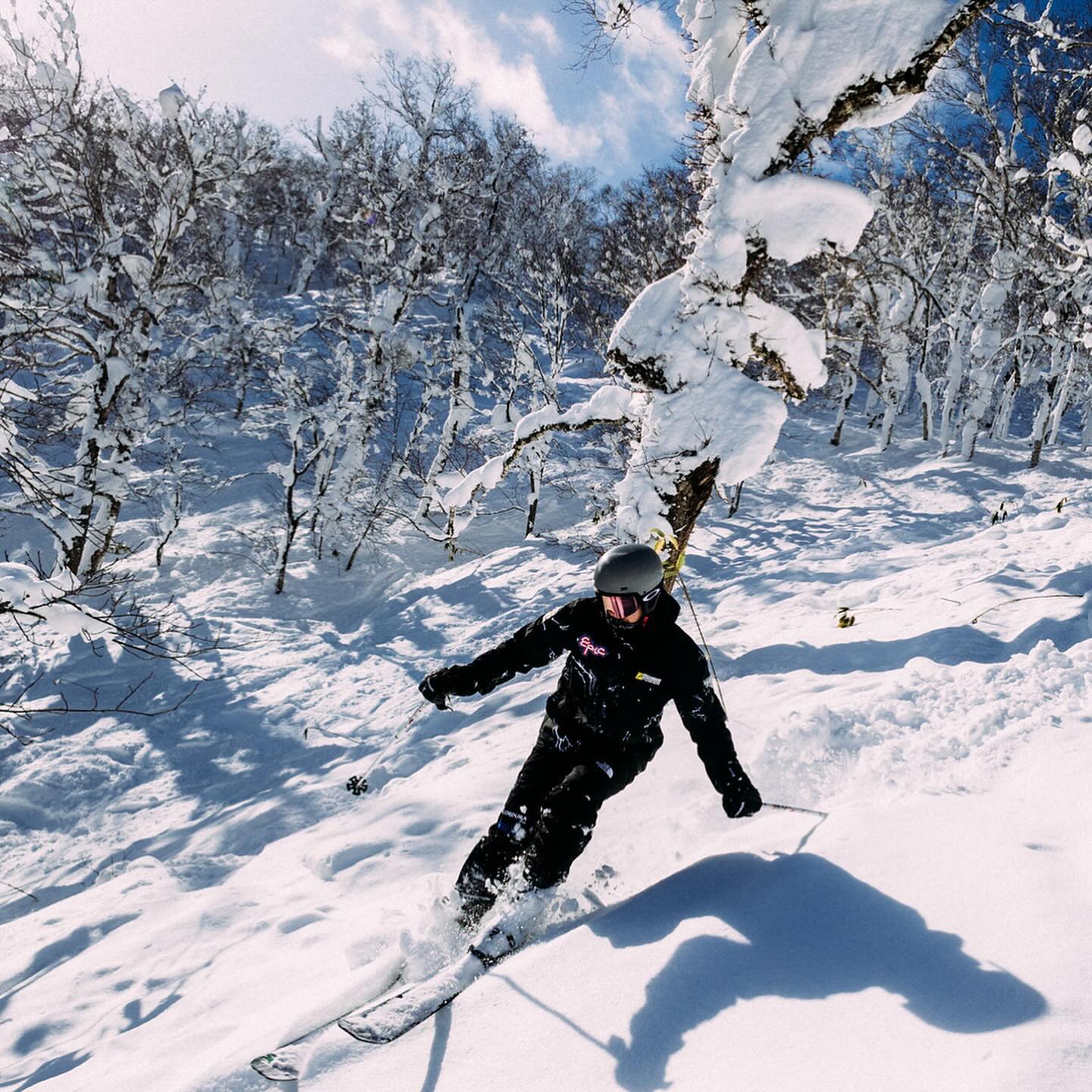 Person skiing down a snow-covered slope surrounded by snow-covered trees on a sunny day, wearing black ski suit and helmet.