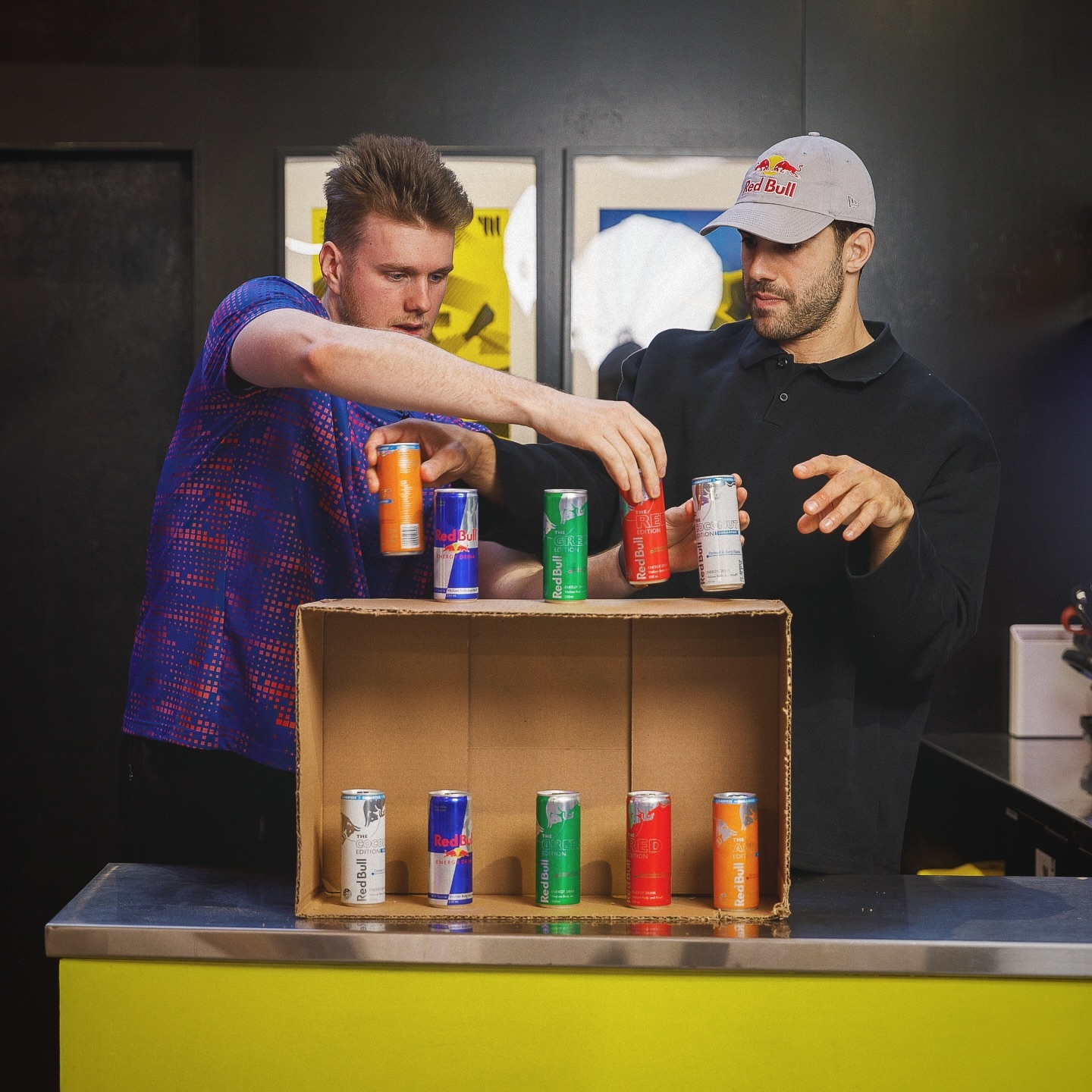 Two adult men arrange Red Bull cans in a cardboard box on a yellow table in an indoor setting with gray and black background.