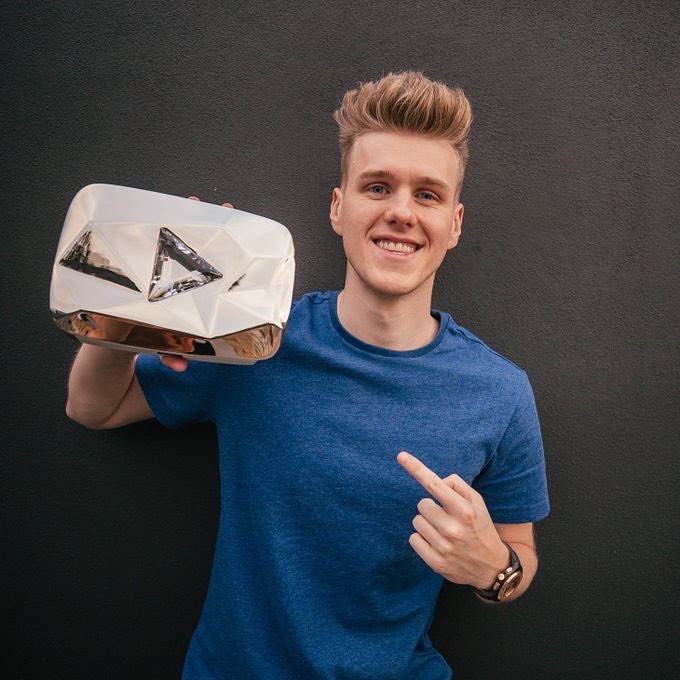 Smiling adult male holds a diamond play button award in front of a dark gray background. He wears a blue t-shirt and a wrist watch.