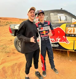 Two men posing next to a yellow racing truck in a sandy desert landscape under a clear sky.