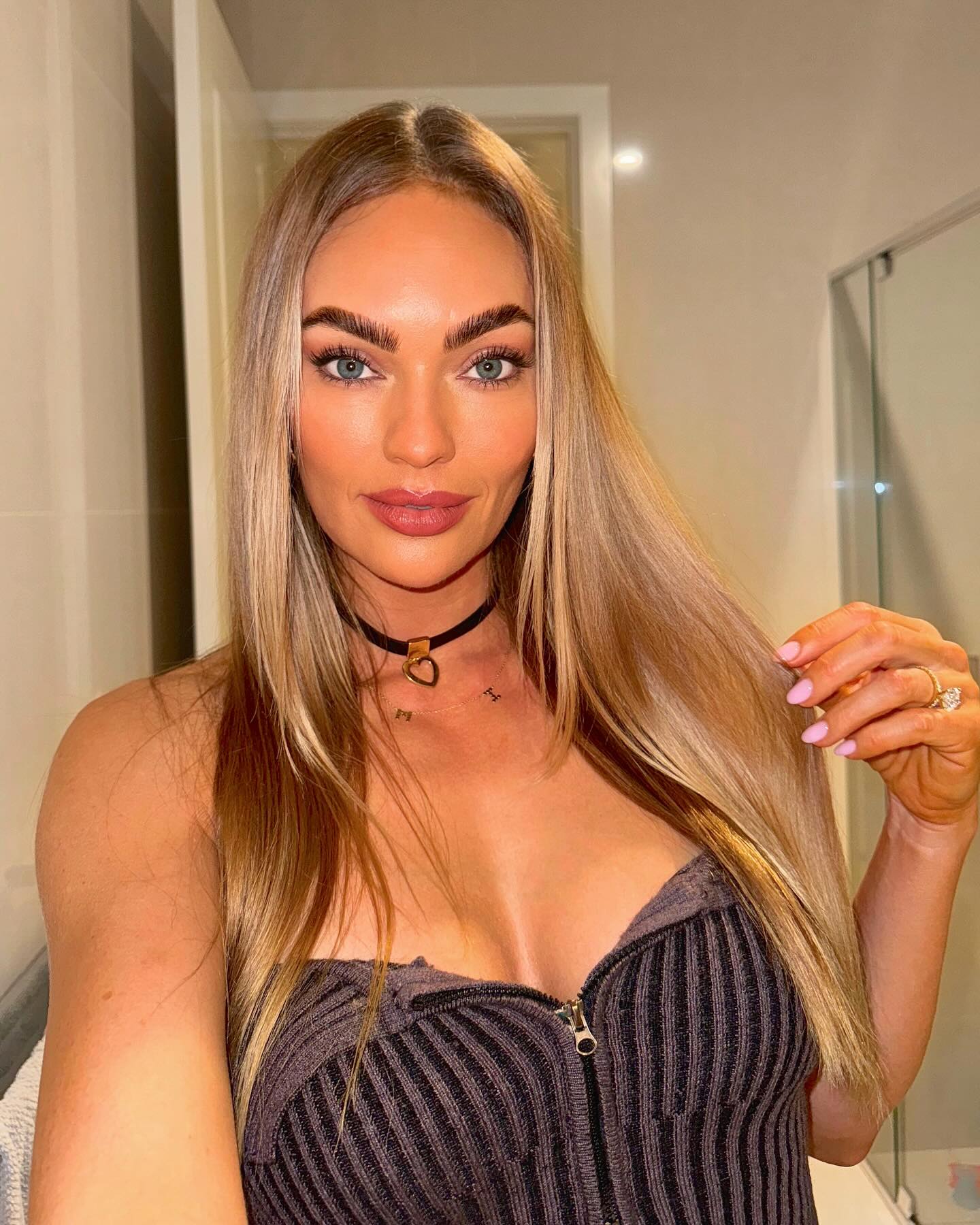 A woman with long blonde hair, wearing a black choker necklace and a striped top, poses indoors in a well-lit setting.