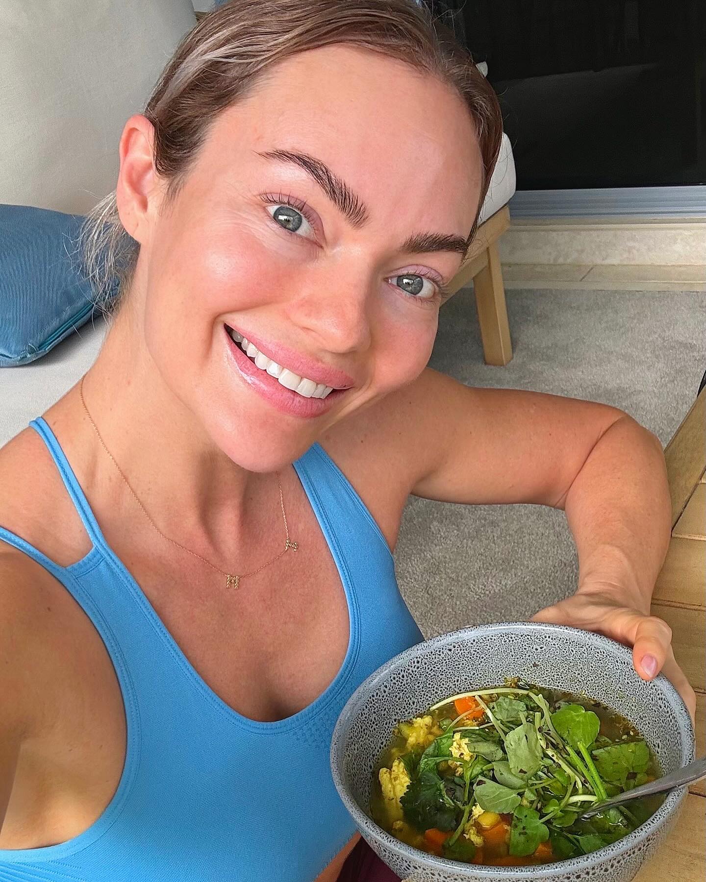Adult woman smiles while holding a bowl of leafy green soup indoors, wearing a blue top with a gold necklace.