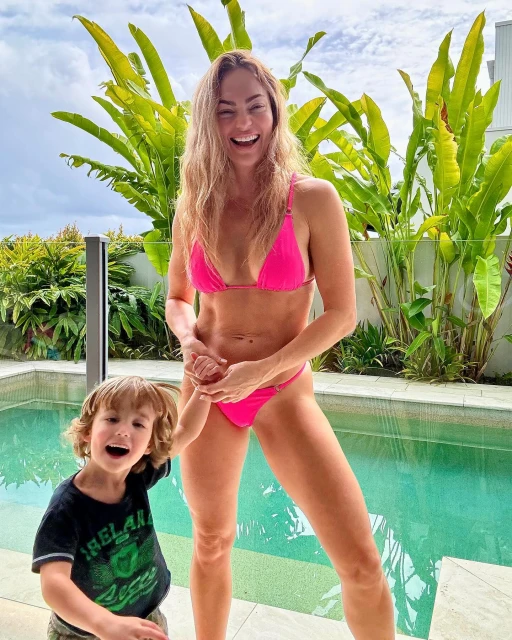 Woman and child pose outdoors by a pool on a sunny day. The woman wears a bright pink bikini; the child wears a dark green tee.