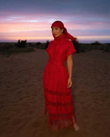 Woman in a vibrant red dress and matching headscarf standing on a sandy desert path at dusk under a purple sunset sky.