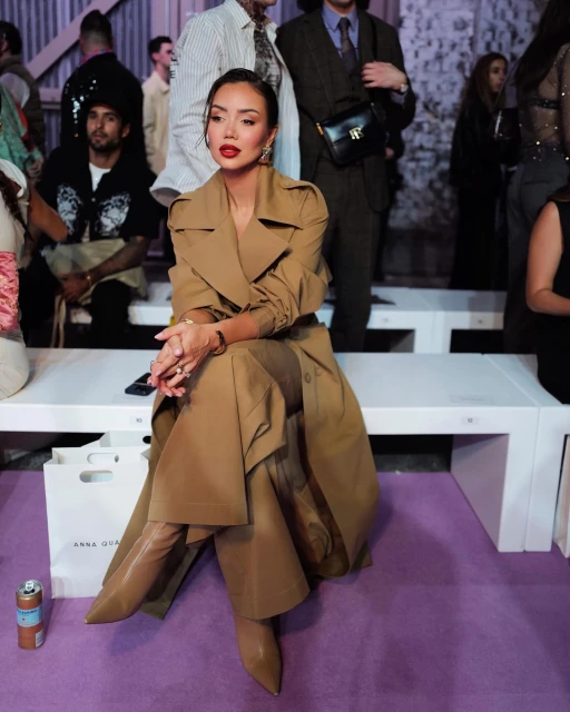Woman in a trench coat and boots sitting on a bench at a fashion show, with other attendees in the background. Editorial fashion shot.