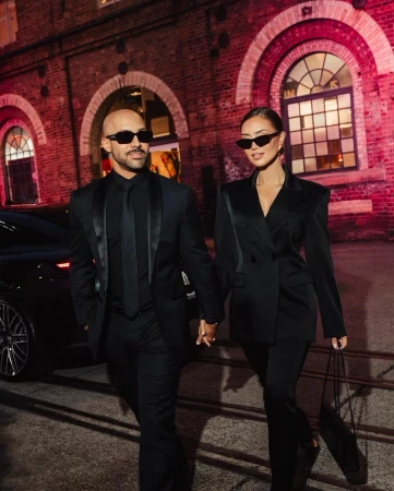 Man and woman holding hands walking outdoors at night wearing matching black suits and sunglasses