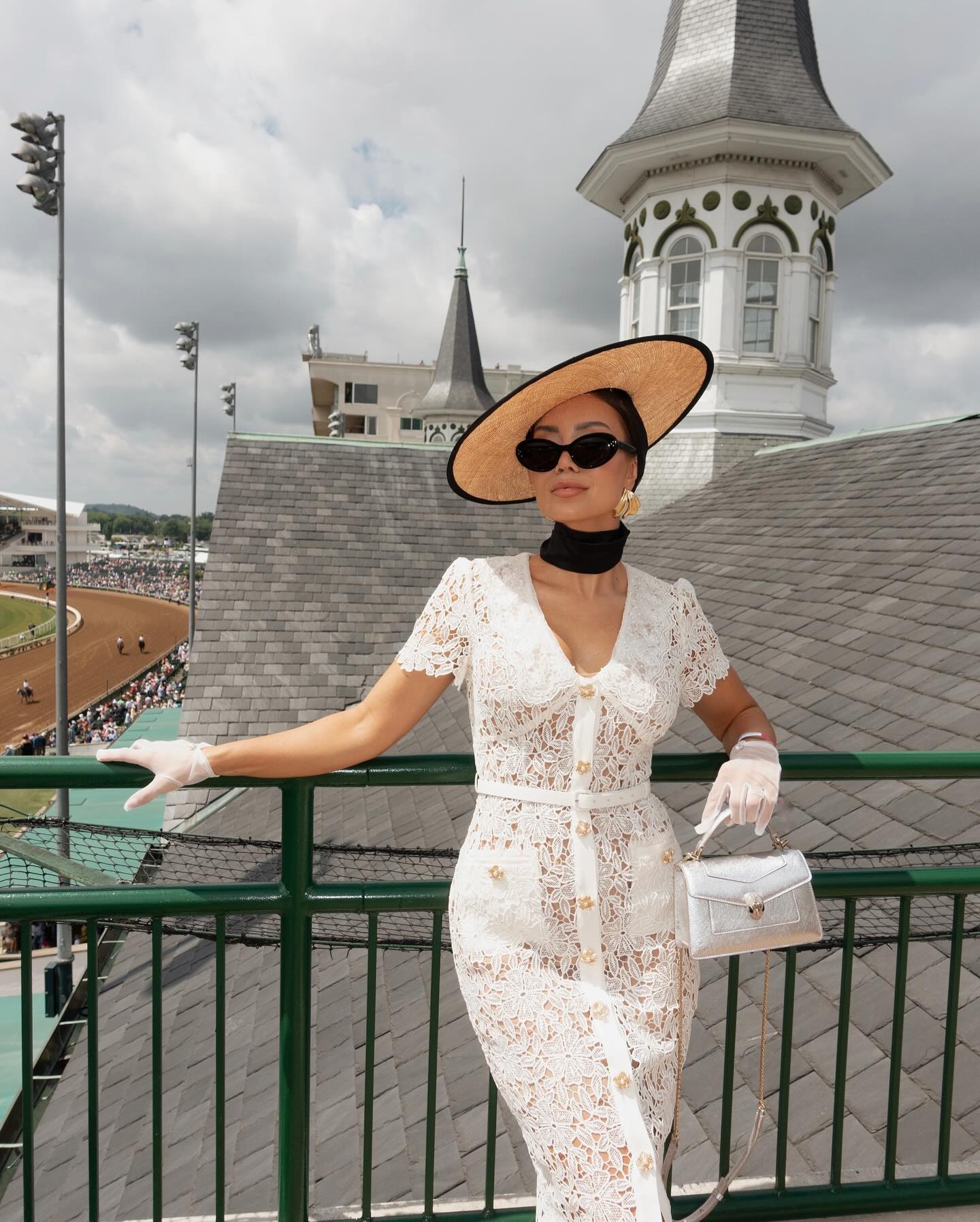 Woman in white lace dress, straw hat, and sunglasses at a horse race event, posing on a balcony overlooking the racetrack.