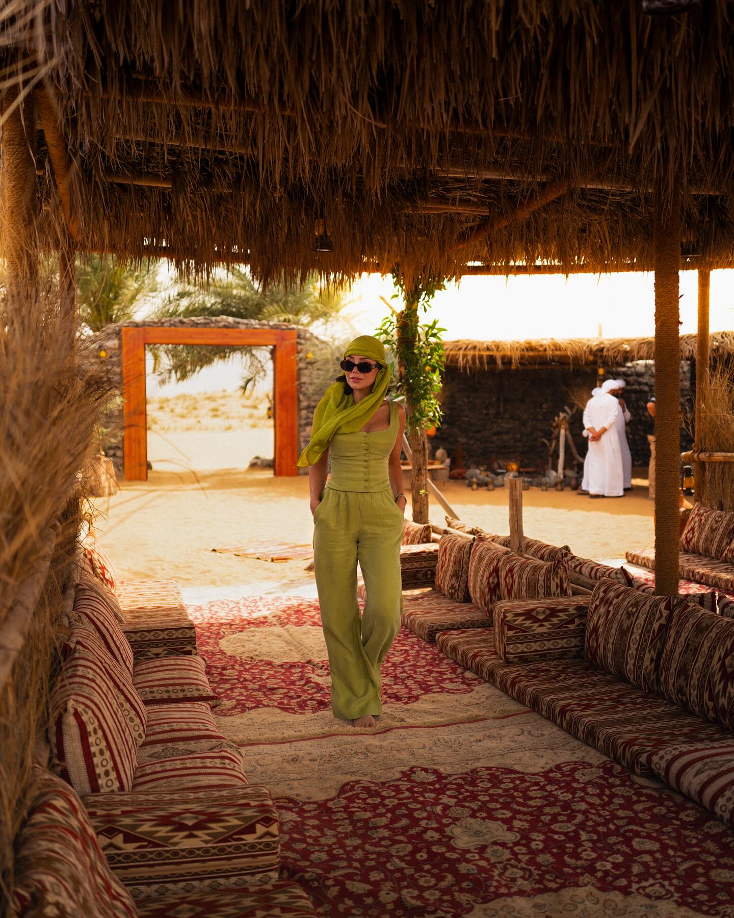 A stylish adult female in green outfit walks on a patterned carpet under a thatched roof structure in an outdoor desert setting.