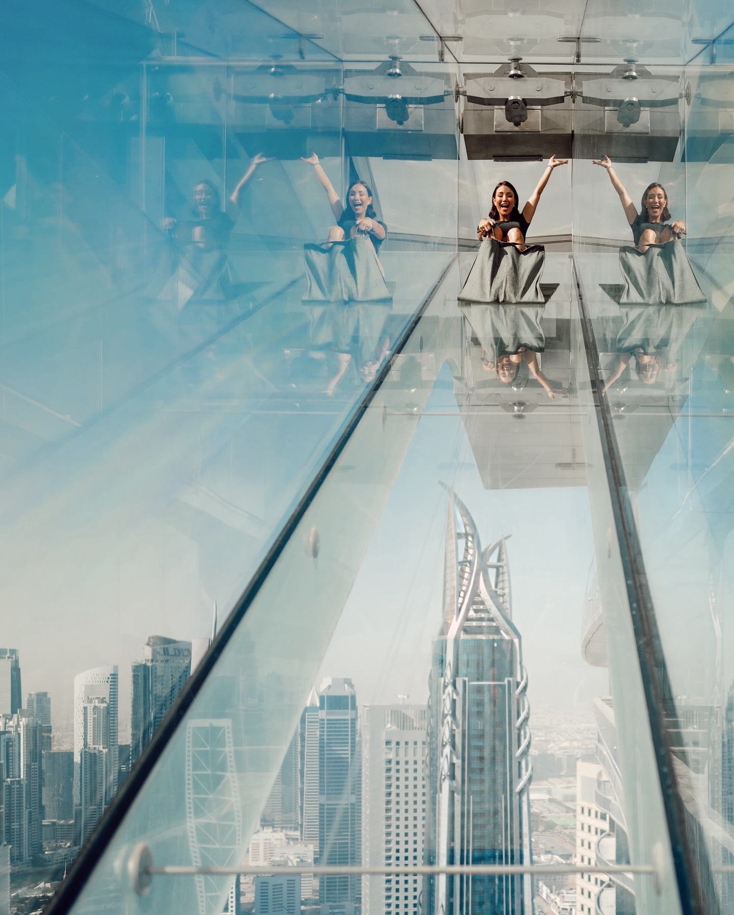 Three women sit on the transparent floor of a high-rise structure, arms raised, showcasing an expansive city view in the background.