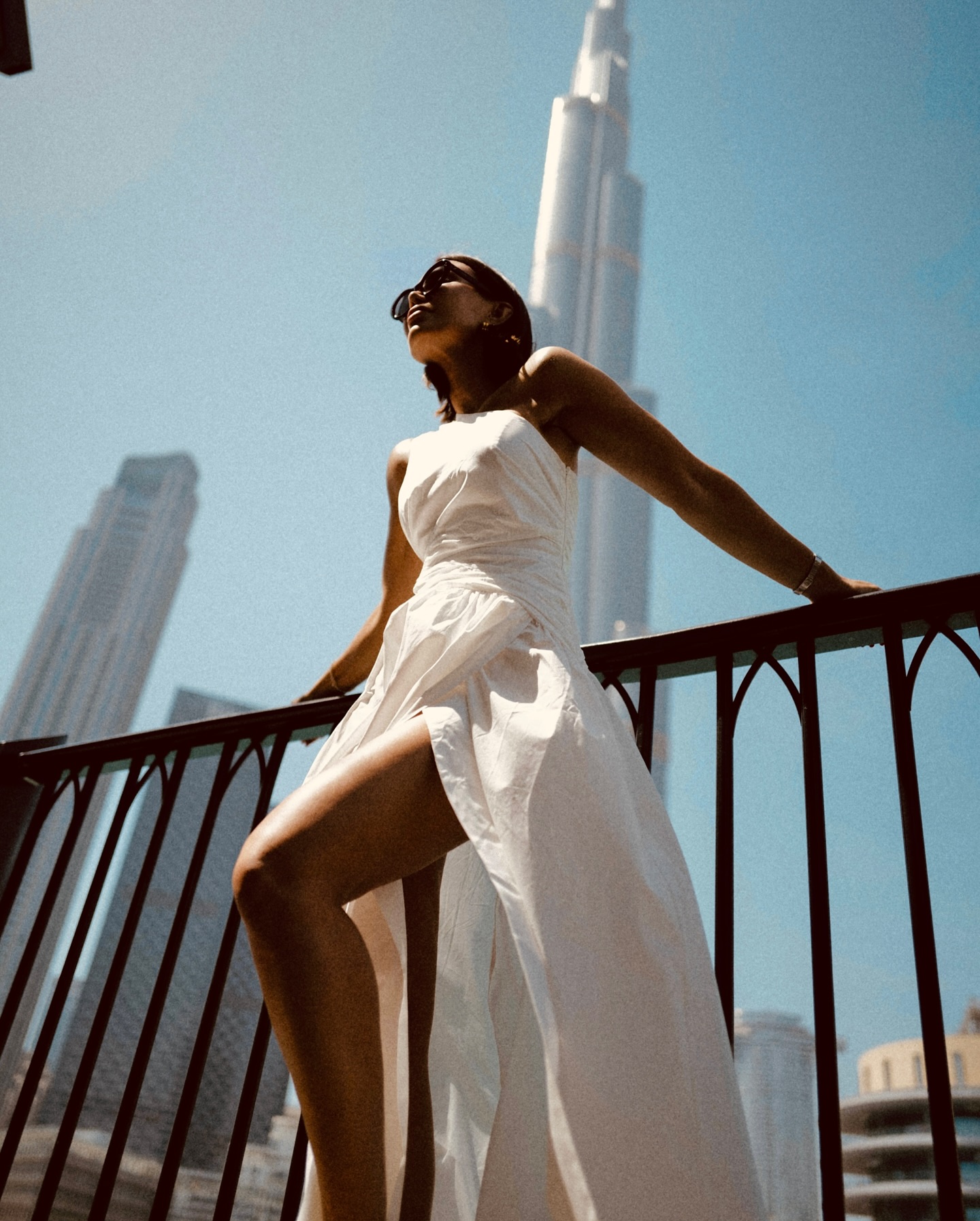Adult female poses in a long, white dress on a balcony with high-rise city buildings in the background on a sunny day.