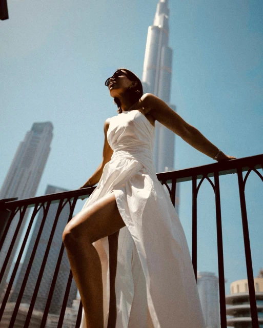Adult female poses in a long, white dress on a balcony with high-rise city buildings in the background on a sunny day.