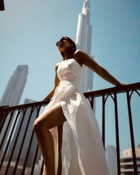 Woman in a white dress leaning on a railing against a towering skyscraper and clear blue sky background.