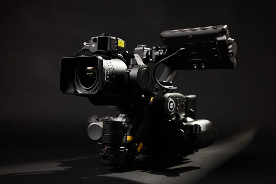 Close-up shot of a professional video camera with a lens hood, featuring a matte black finish against a solid black background.