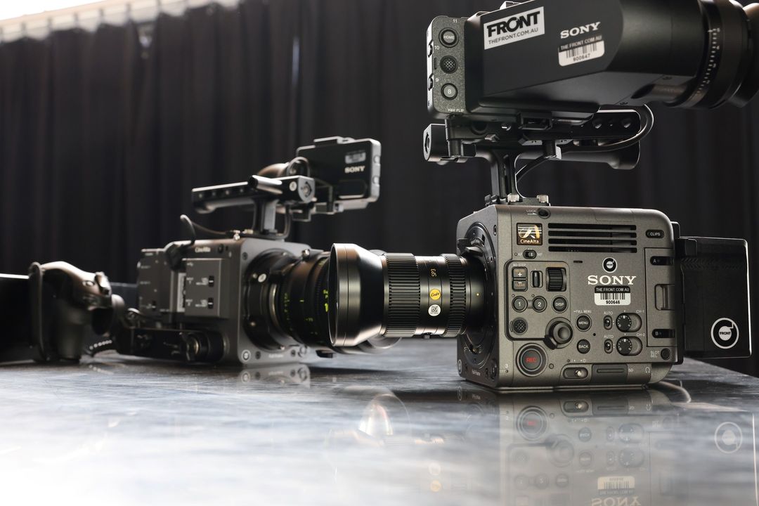 Two Sony professional video cameras with lenses are on a table against a black backdrop, illuminated by studio lighting.