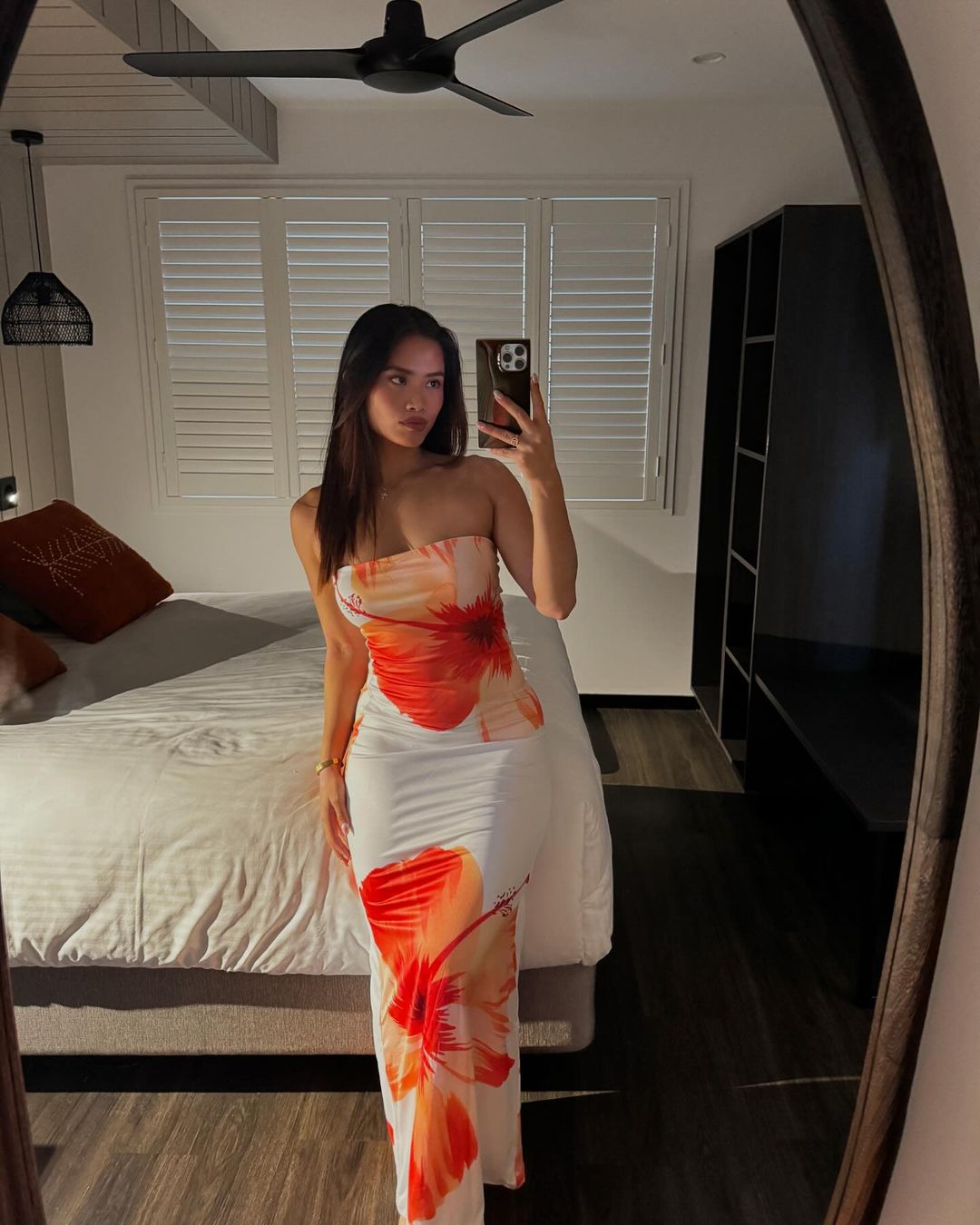 Young woman in floral dress takes mirror selfie in bedroom. Interior view with bed, shutters, neutral colors, and dim lighting.
