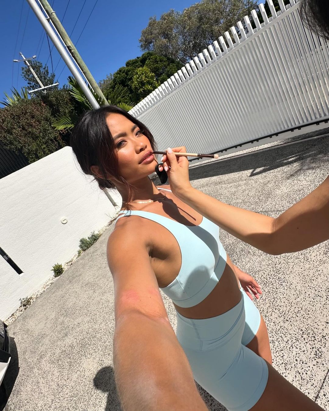 Asian woman having makeup applied outdoors; wearing a matching blue sports bra and shorts set with a fence and blue sky in the background.