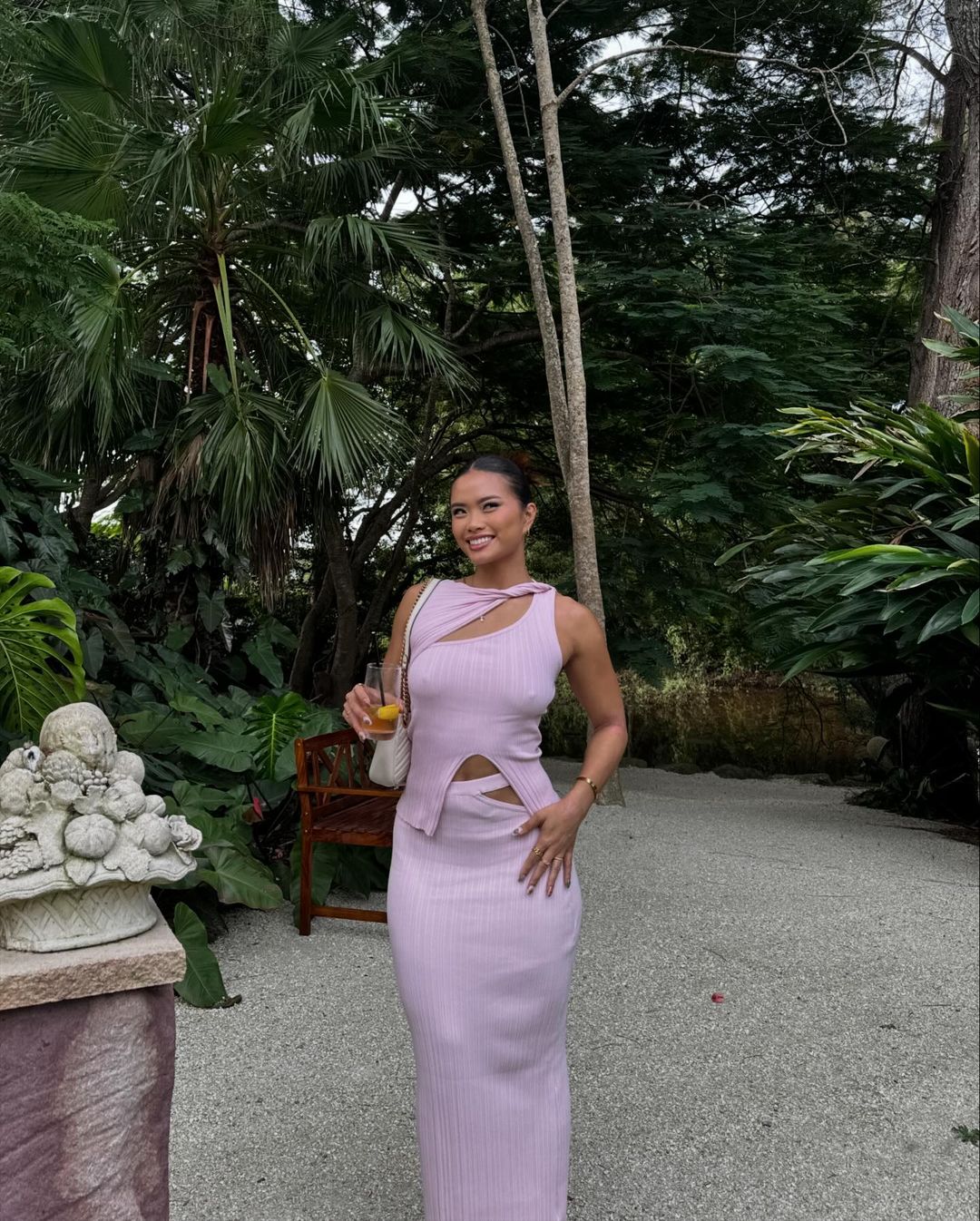 A smiling woman in a pink knit set poses outside surrounded by lush greenery. She is holding a drink and has a purse over her shoulder.