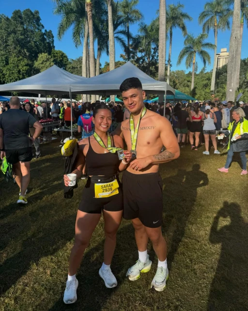 Two runners pose in a grassy park with medals and crowds, wearing athletic wear and sneakers, under palm trees on a sunny day.