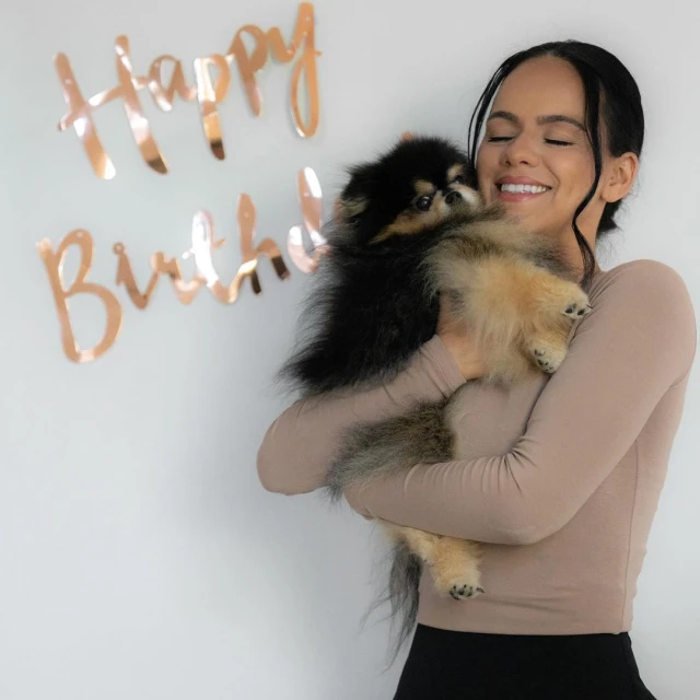 Smiling adult woman embraces fluffy Pomeranian dog in an indoor setting with 'Happy Birthday' in gold foil letters on the wall.