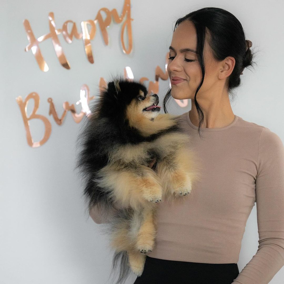 A smiling woman in a beige top holds a Pomeranian dog in an indoor photo studio, with a 'Happy Birthday' sign visible in the background.