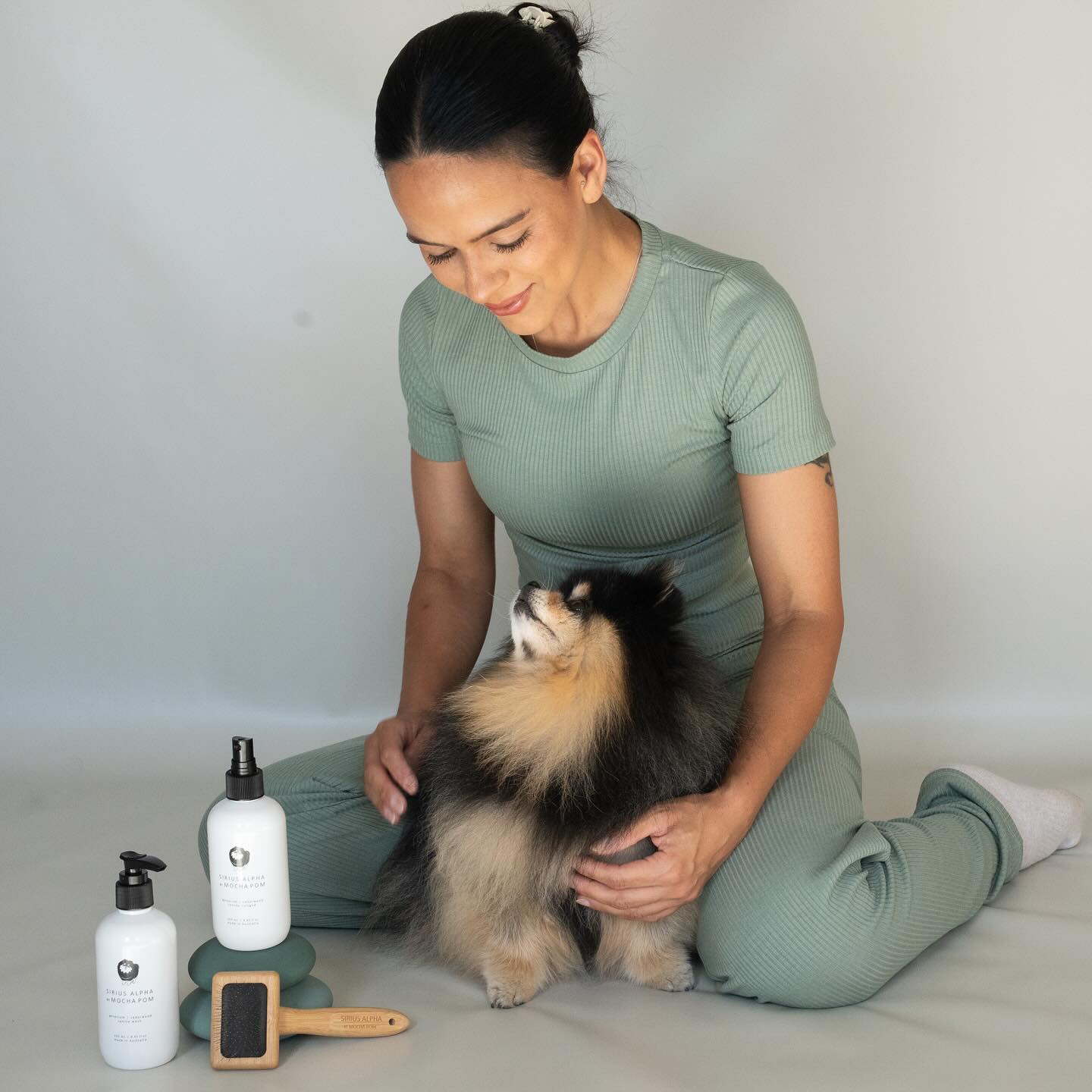 Woman in green athletic wear sits with a Pomeranian dog, posing near grooming products, against a plain studio background, indoors.
