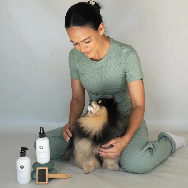 Woman in green athletic wear sits with a Pomeranian dog, posing near grooming products, against a plain studio background, indoors.